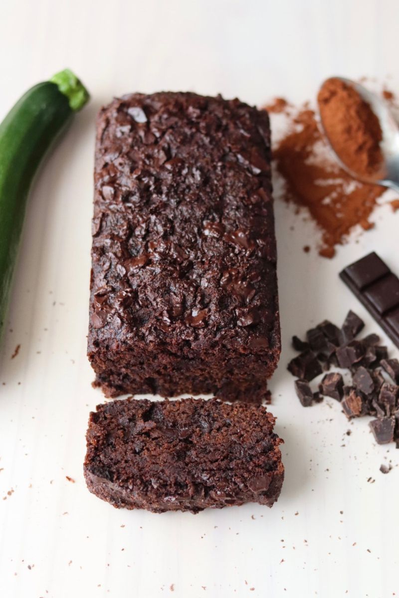 A gluten free zucchini bread with a zucchini on the side and chocolate bar and chips and a spoon full of cacao powder.