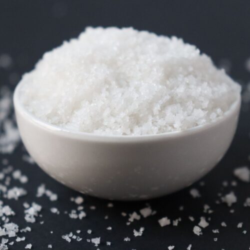 a white bowl filled with fleur de sel on a black table mat, sprinkles of fleur de sel scattered all around