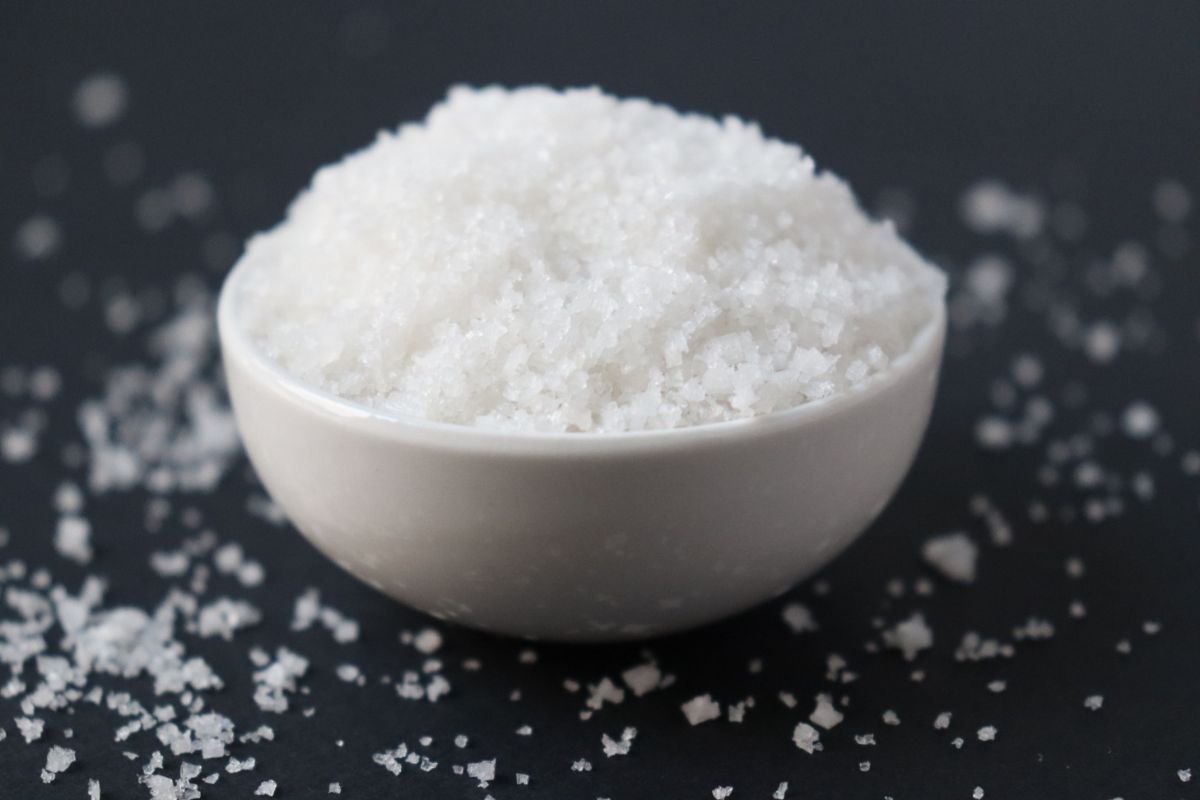 a white bowl filled with fleur de sel on a black table mat, sprinkles of fleur de sel scattered all around