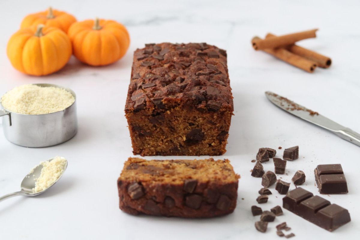 a chocolate chip pumpkin bread with one slice cut off, small pumpkins cinnamon sticks, chocolate chips and almond flour around it to decorate
