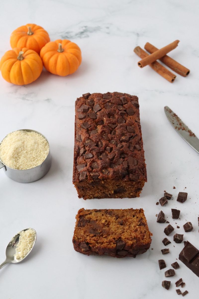 a chocolate chip pumpkin bread with one slice cut off, small pumpkins cinnamon sticks, chocolate chips and almond flour around it to decorate