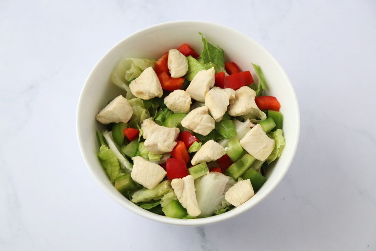 Salade, cubes de poulet et poivron rouge et vert coupé en morceaux dans un saladier blanc.