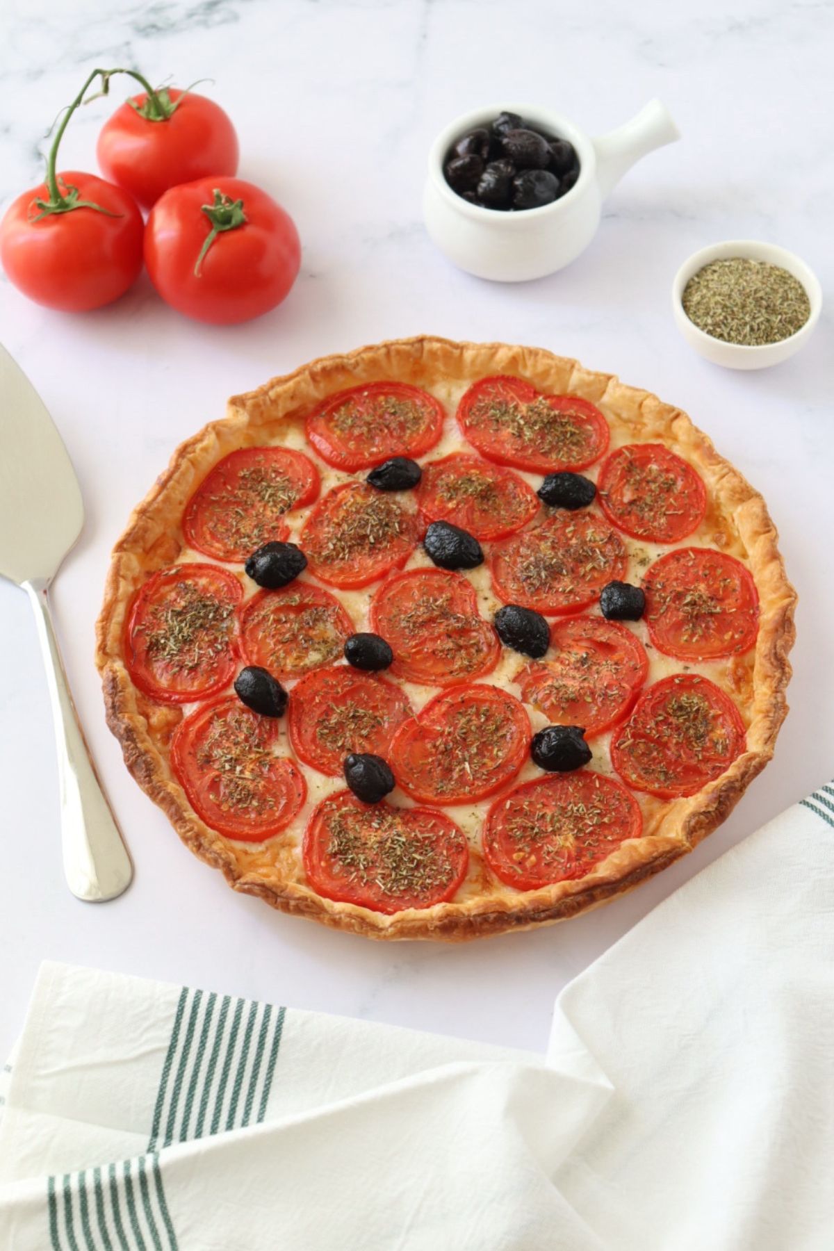 Une tarte à la tomate entourées de 3 tomates, un petit bol d'herbes de Provence, un bol d'olives noires, une pelle à tarte et un torchon.