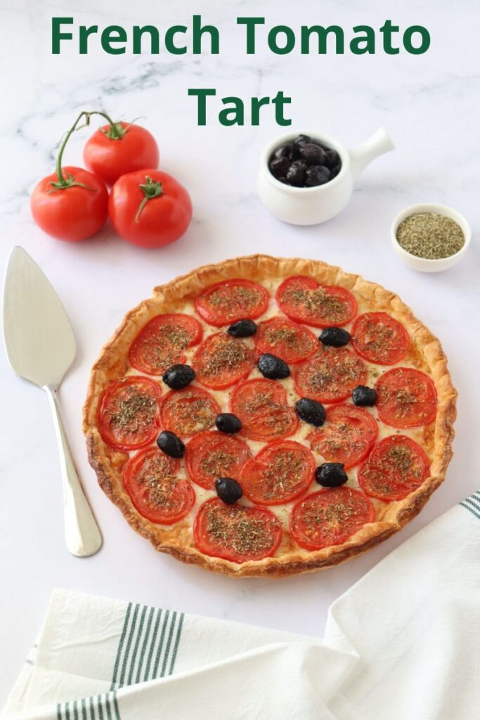 A tomato tart surrounded with 3 tomatoes, a small pot of herbs de Provence, a pot of black olives, a pie server and a kitchen cloth and a text overlay with the title.