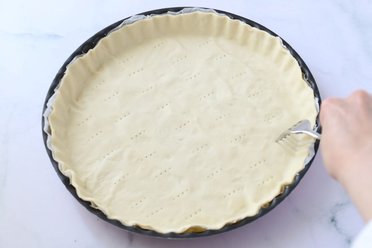 Une pâte feuilletée dans un moule à tarte et une fourchette en train de piquer la pâte.