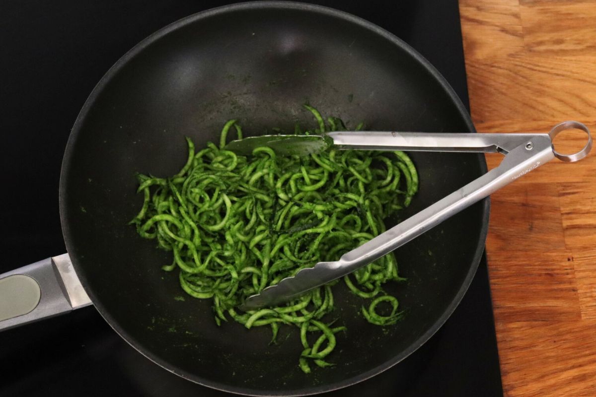 Zucchini noodles coated with pesto in a wok with kitchen tongs on the side.