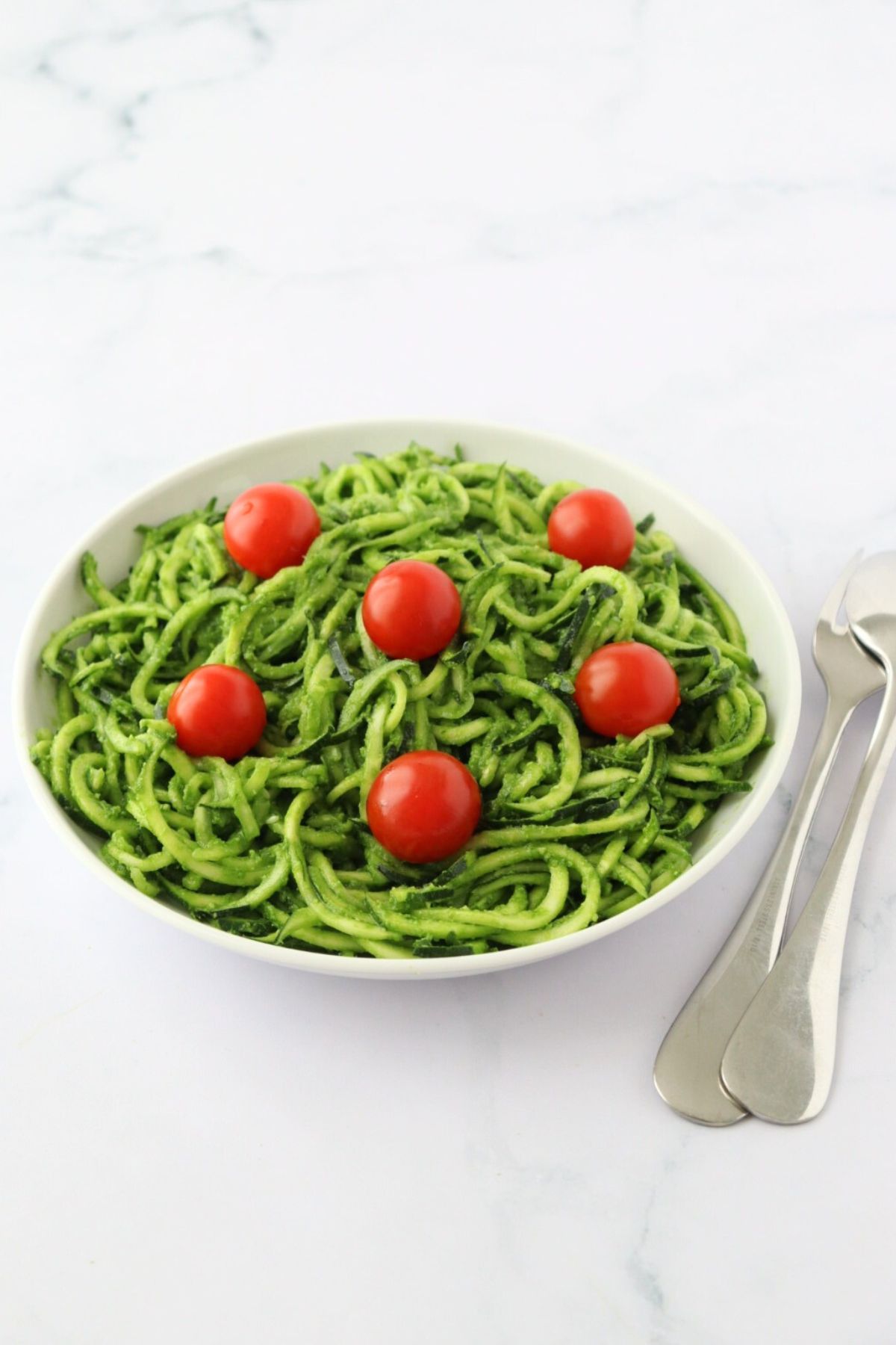 A plate full of zucchini noodles coated with pesto and some cherry tomatoes on top, a spoon and a fork on the side.