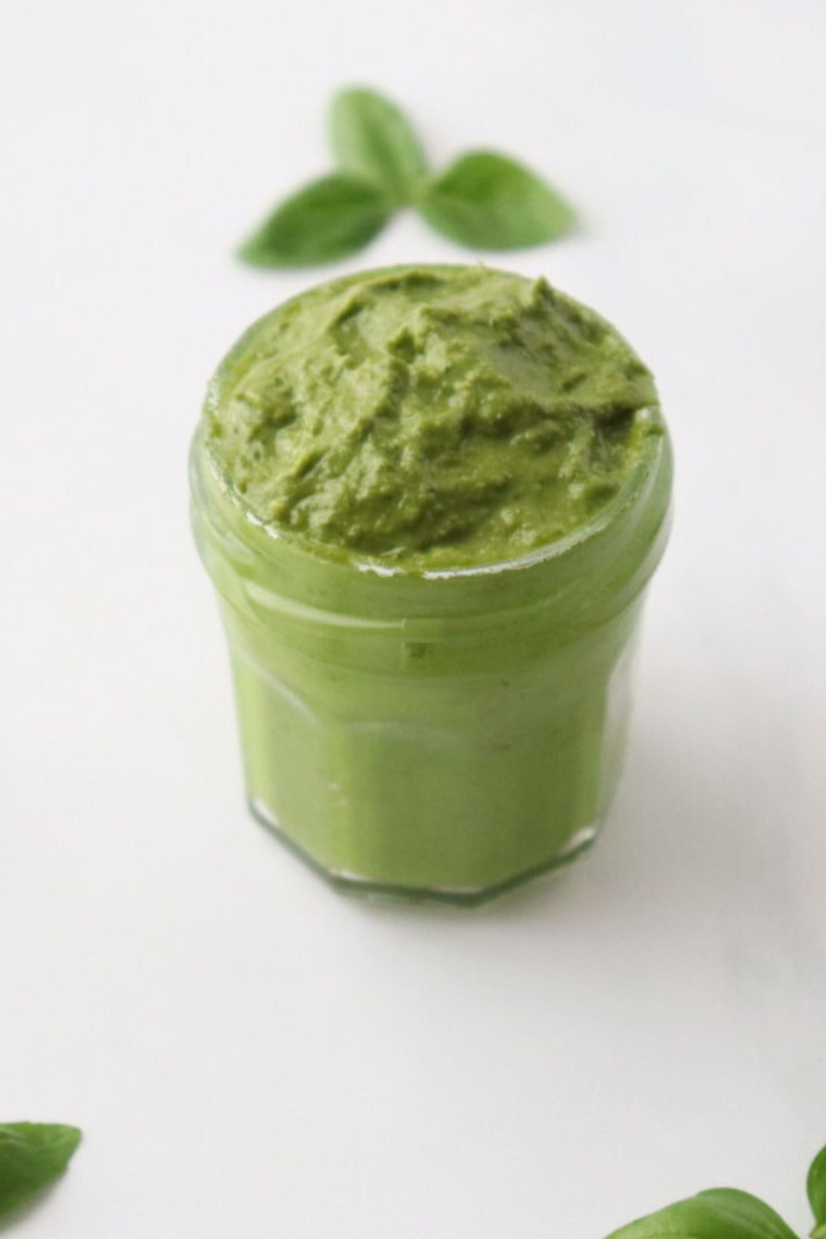 A jar full of basil pesto without pine nuts, basil leaves around it.