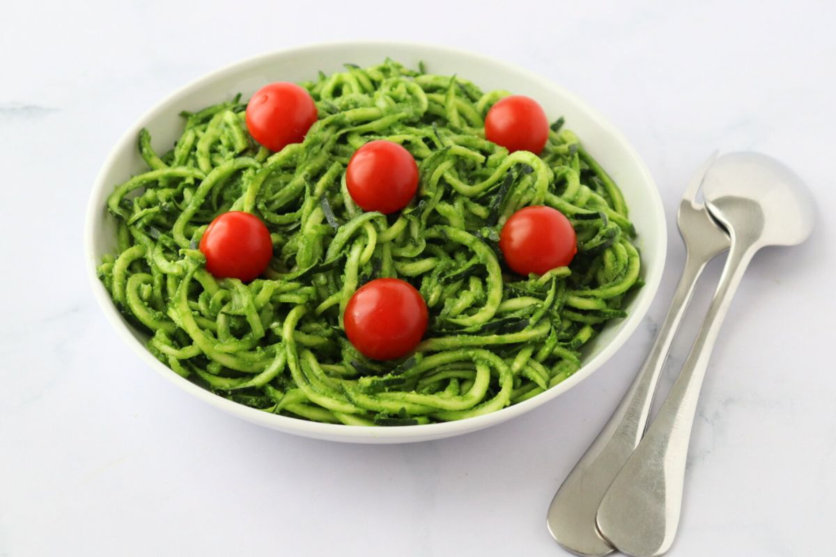 A plate full of zucchini noodles coated with pesto and some cherry tomatoes on top, a spoon and a fork on the side.