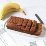 Un banana bread tranché sur un plat blanc, des bananes et un couteau à côté.