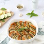 Un tian de légumes sans aubergines et sans tomates entouré de rondelles de courgettes sur une planche, des herbes de Provence, des gousses d'ail et un huilier.
