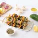 A plate full of chicken kabobs surrounded by slices of lemon, red onion, green zucchini, yellow zucchini, olive oil, herbs de Provence and white mushrooms.