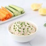 A bowl full of tuna dip sprinkled with chopped chives, some vegetable sticks, lemon quarters and chives on the side.