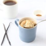 A vanilla mug cake with some vanilla beans on the side, a cup of tea and a small bowl of cinnamon in the background.