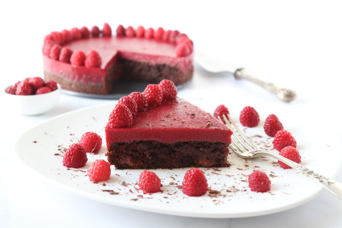 A slice of chocolate raspberry cake on a plate decorated with raspberries and chocolate shavings along with the whole cake in the background.