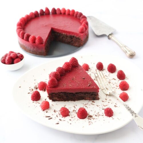 A slice of chocolate raspberry cake on a plate decorated with raspberries and chocolate shavings along with the whole cake in the background.
