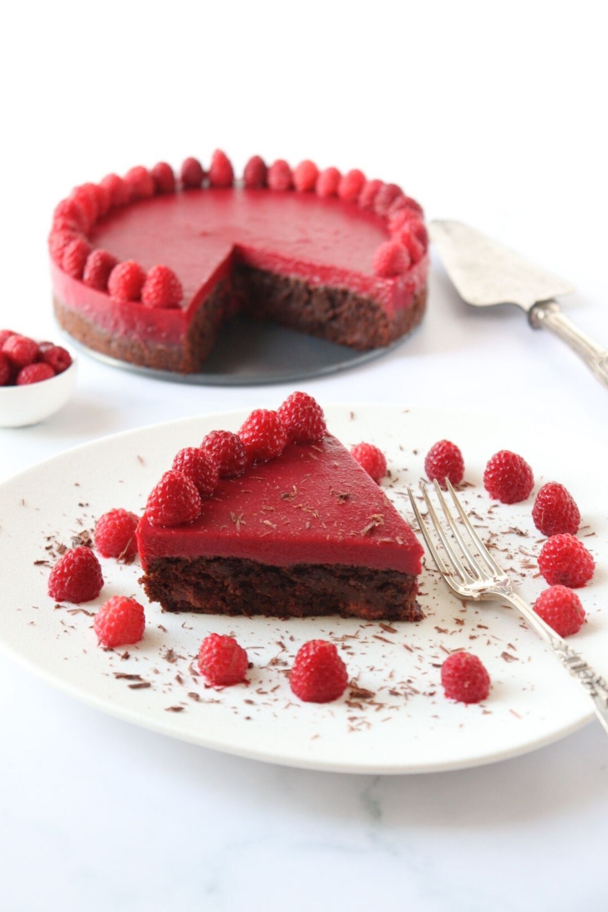 A slice of chocolate raspberry cake on a plate decorated with raspberries and chocolate shavings along with the whole cake in the background.