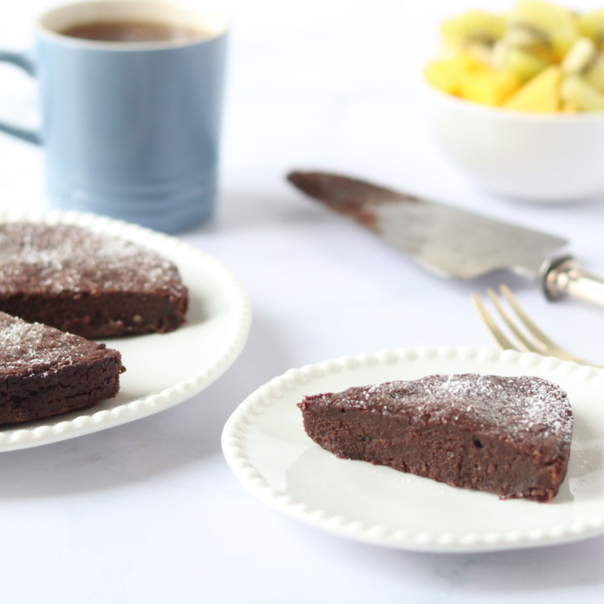 A slice of butternut squash chocolate cake on a plate next to the rest of the whole cake, a cup of tea and a bowl of fruit salad in the background.