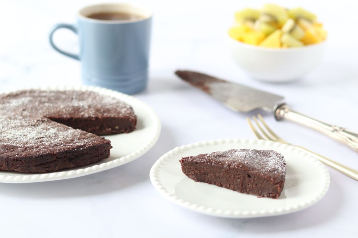 A slice of butternut squash chocolate cake on a plate next to the rest of the whole cake, a cup of tea and a bowl of fruit salad in the background.