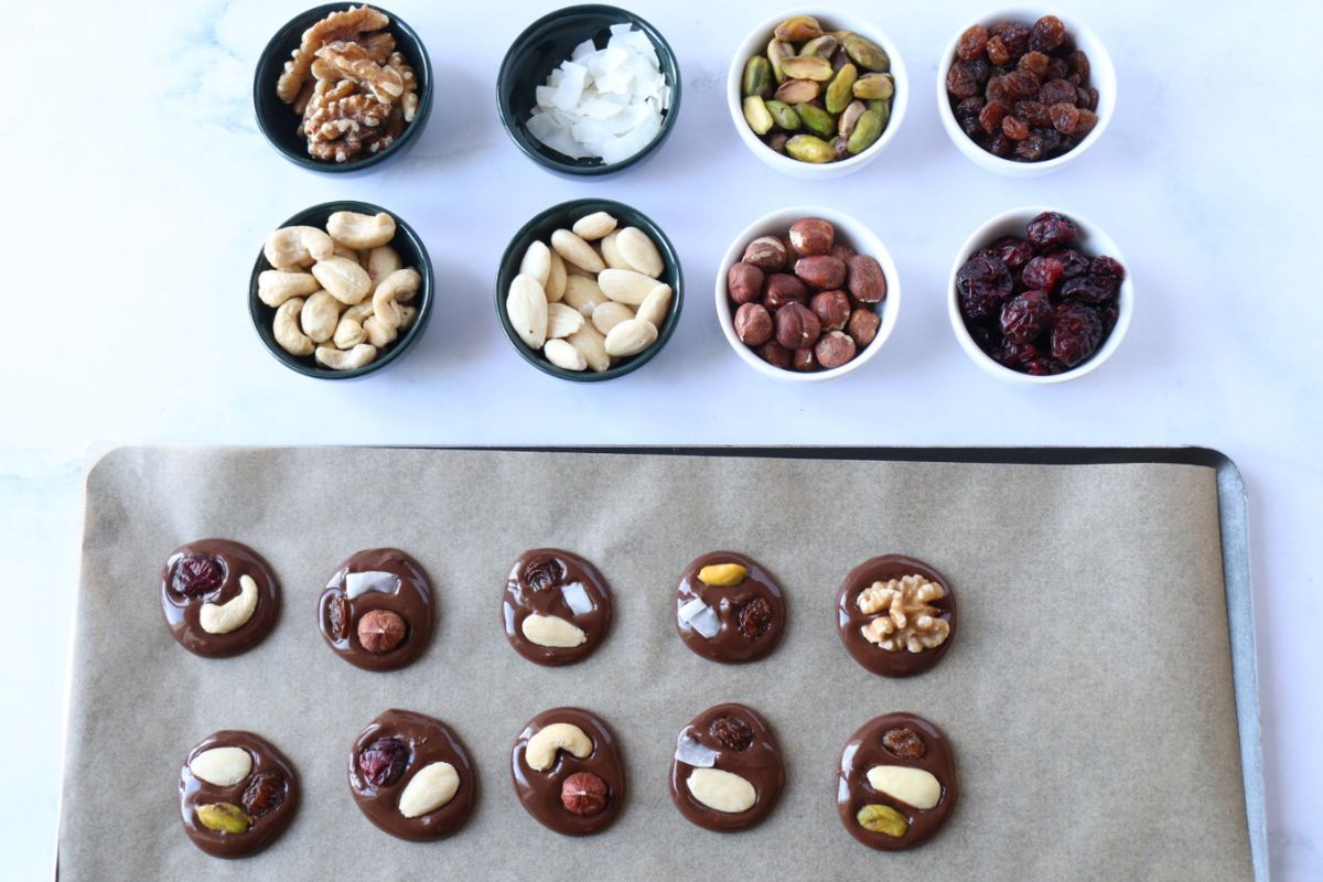 Chocolate disks covered with nuts and dried fruits on a lined baking tray along with all the nuts and dried fruits prepared in separate bowls.