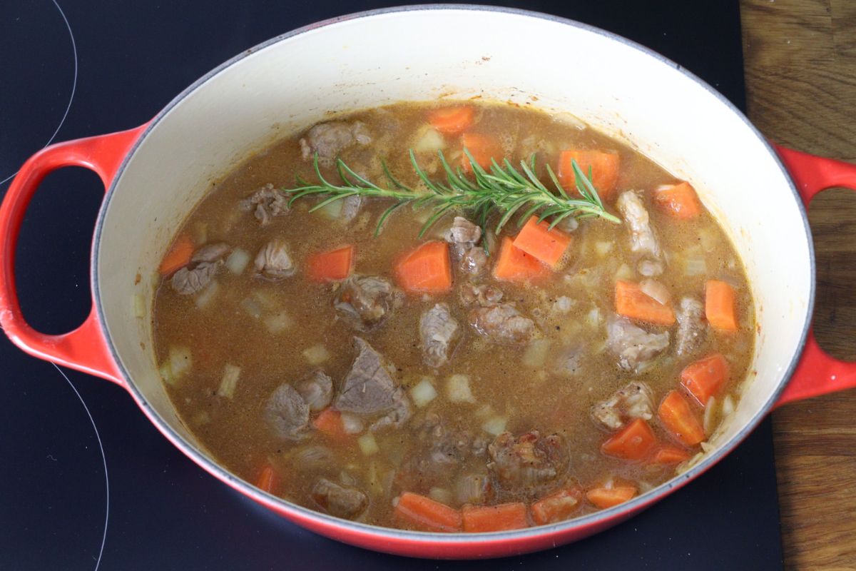 Lamb meat cooking in a Dutch oven with carrots, onion, garlic and arrowroot and tomato paste mixed in it topped with water and a sprig of rosemary.
